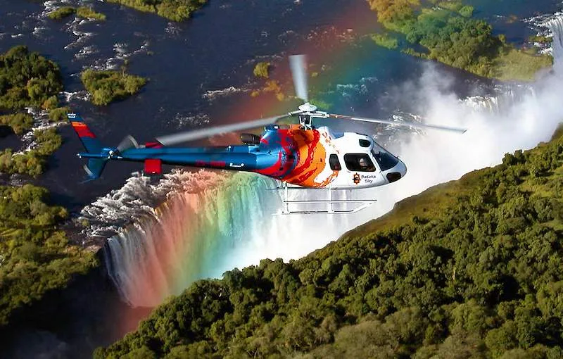 A helicopter flies above Victoria Falls, with a rainbow rising from the spray.