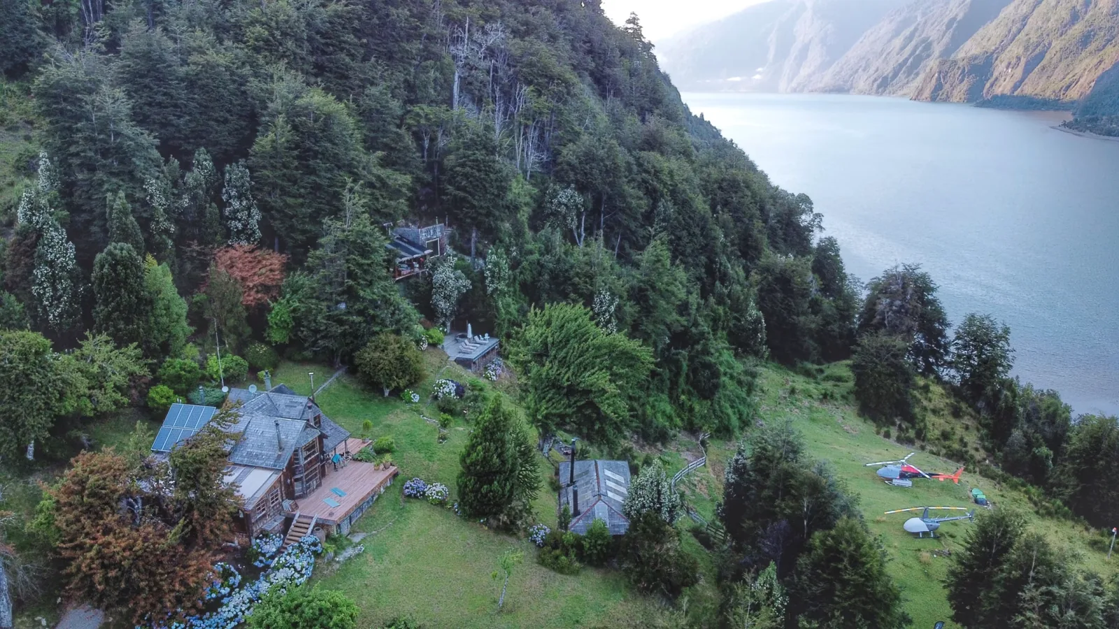 Wooden lodge buildings sit among forested hills above a river.