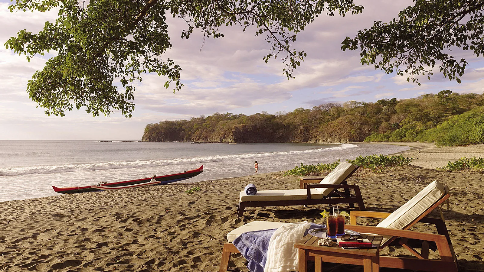 Sun loungers and a kayak rest on a sandy cove edged by tropical trees.