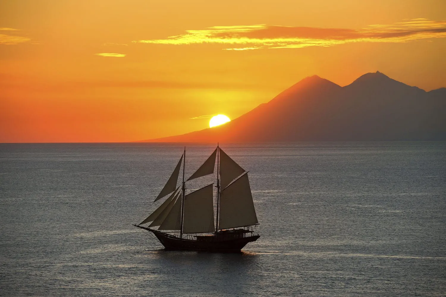 A traditional Indonesian Phinisi yacht sailing past island silhouettes at sunset.