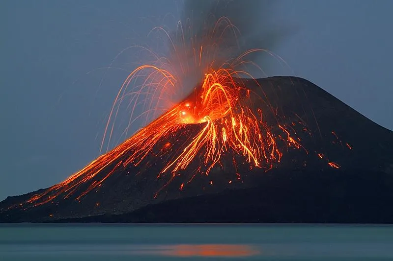 A volcanic island glowing at sunset above the sea, with a sailing boat in the foreground.