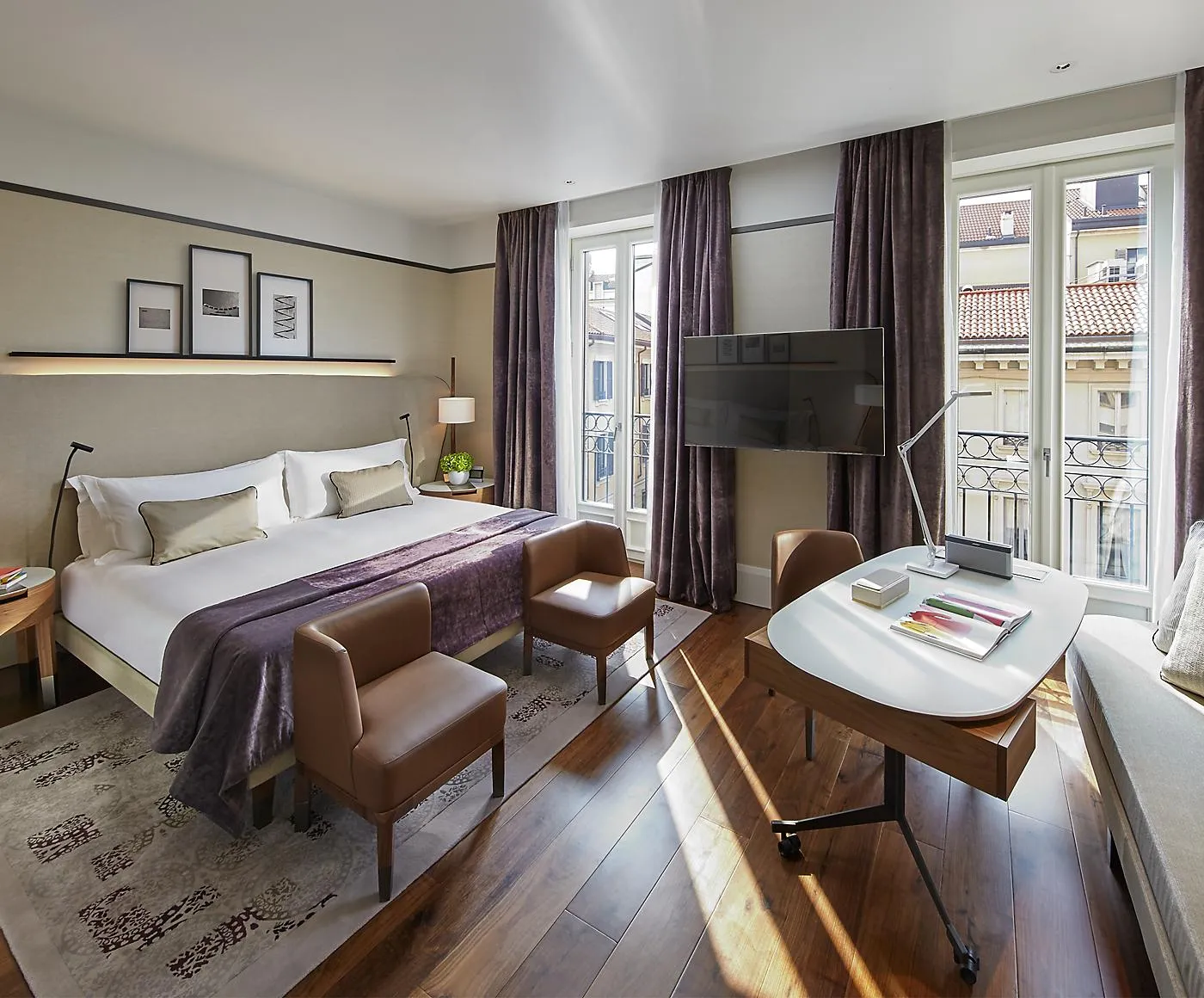 A bright hotel bedroom with a white bed, seating area, and balcony doors.