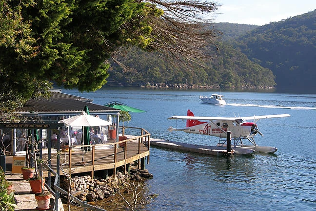 A white and red seaplane moored beside a waterfront restaurant on a wooded inlet.