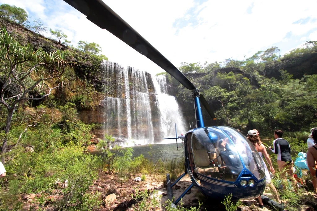 A helicopter on the ground beside a waterfall surrounded by green vegetation.