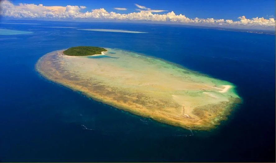 A small sandy island and shallow reef surrounded by deep blue sea.