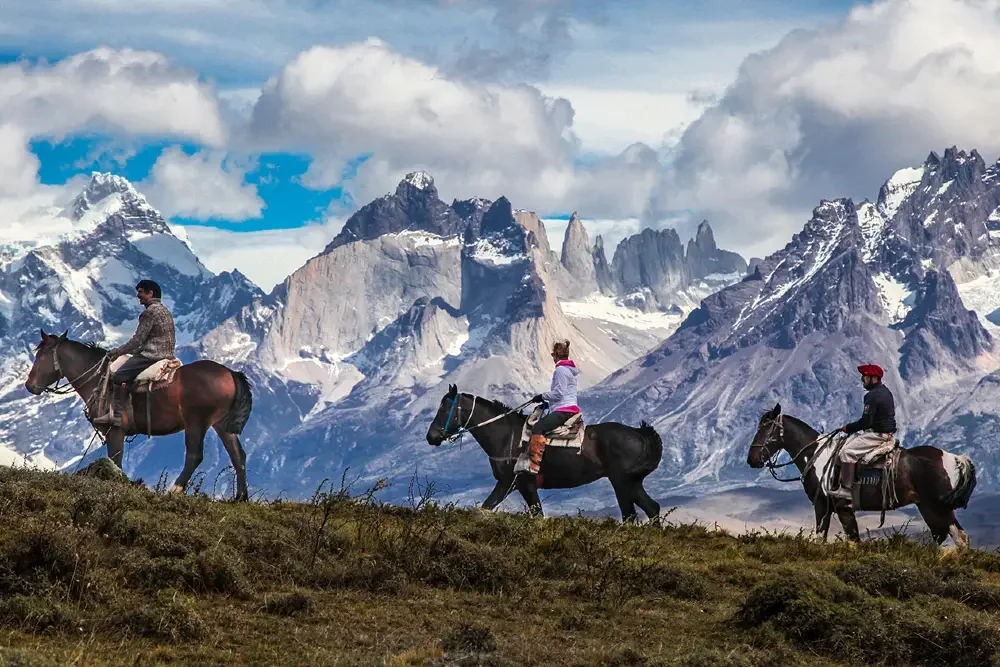 Horse riders crossing grassland beneath rugged Patagonian mountains.