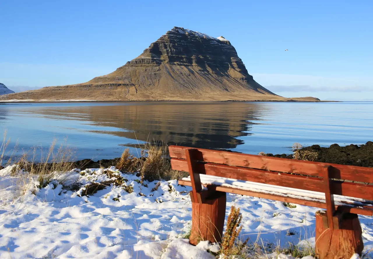 A snow-dusted bench overlooking calm water and a steep, isolated mountain.
