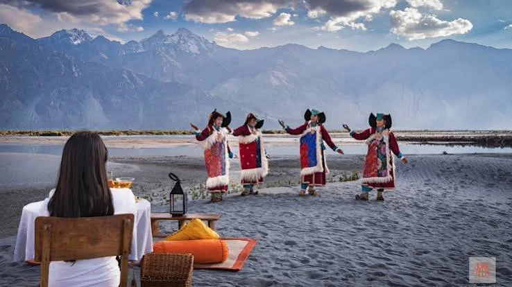 Dancers performing on a sandy plain with mountains behind them and seated guests in the foreground.