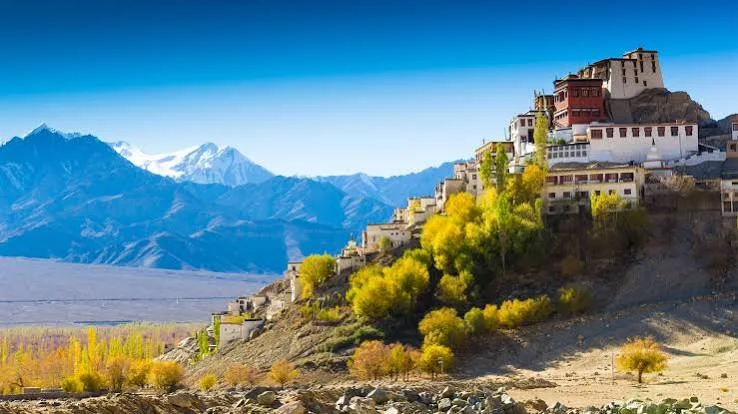 A monastery on a rocky hillside with autumn trees and snowy mountains beyond.