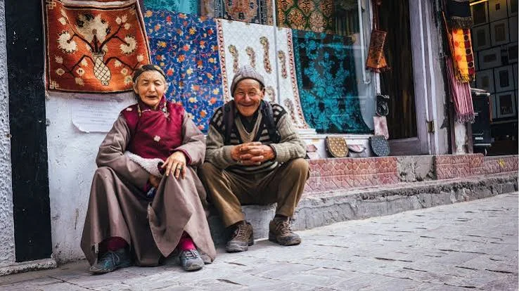 Two people seated outside a shop displaying patterned textiles and colourful crafts.