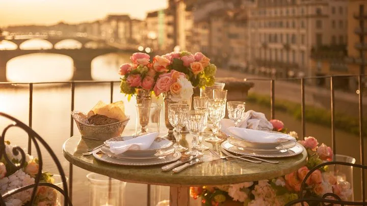 A dining table with flowers on a terrace overlooking Florence's river and bridges at sunset.