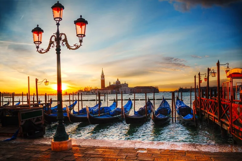 Gondolas moored beside ornate street lamps on Venice's waterfront at sunset.