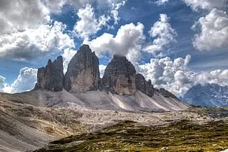 Three jagged Dolomite peaks rising above a rocky plateau beneath clouds.