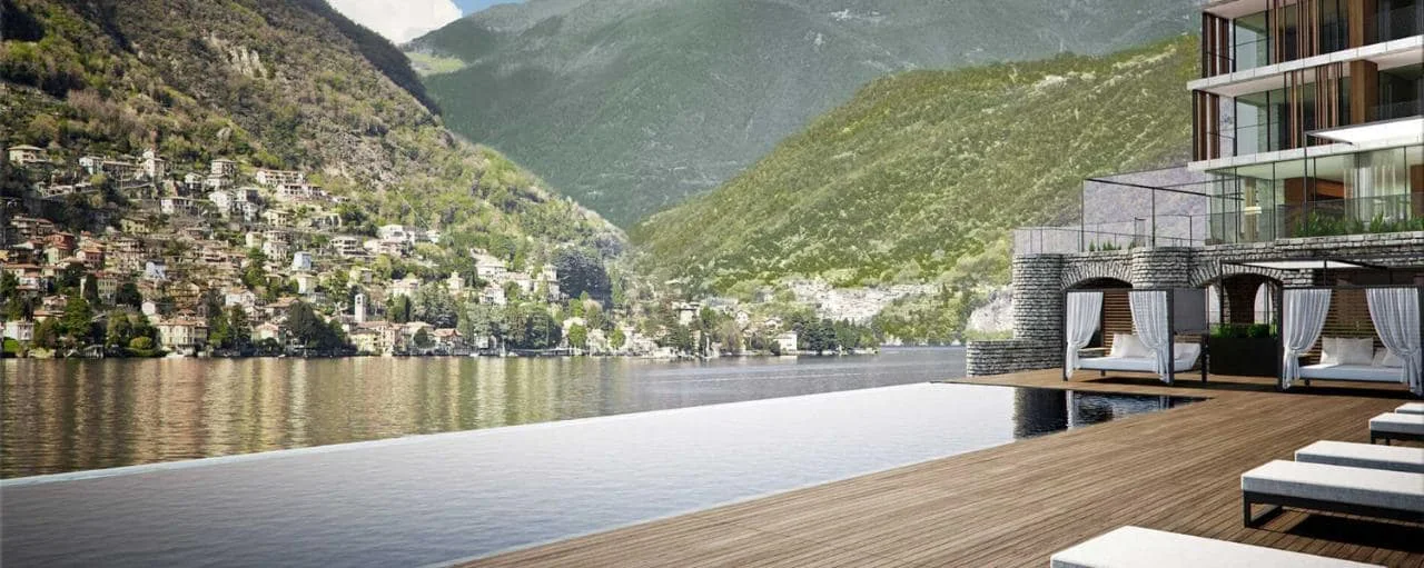 A modern lakeside hotel's infinity pool overlooking water and green mountains.