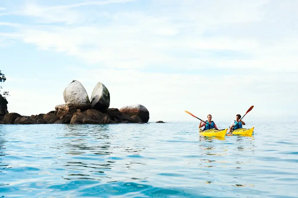 Two people kayaking near a split rock in clear coastal water.