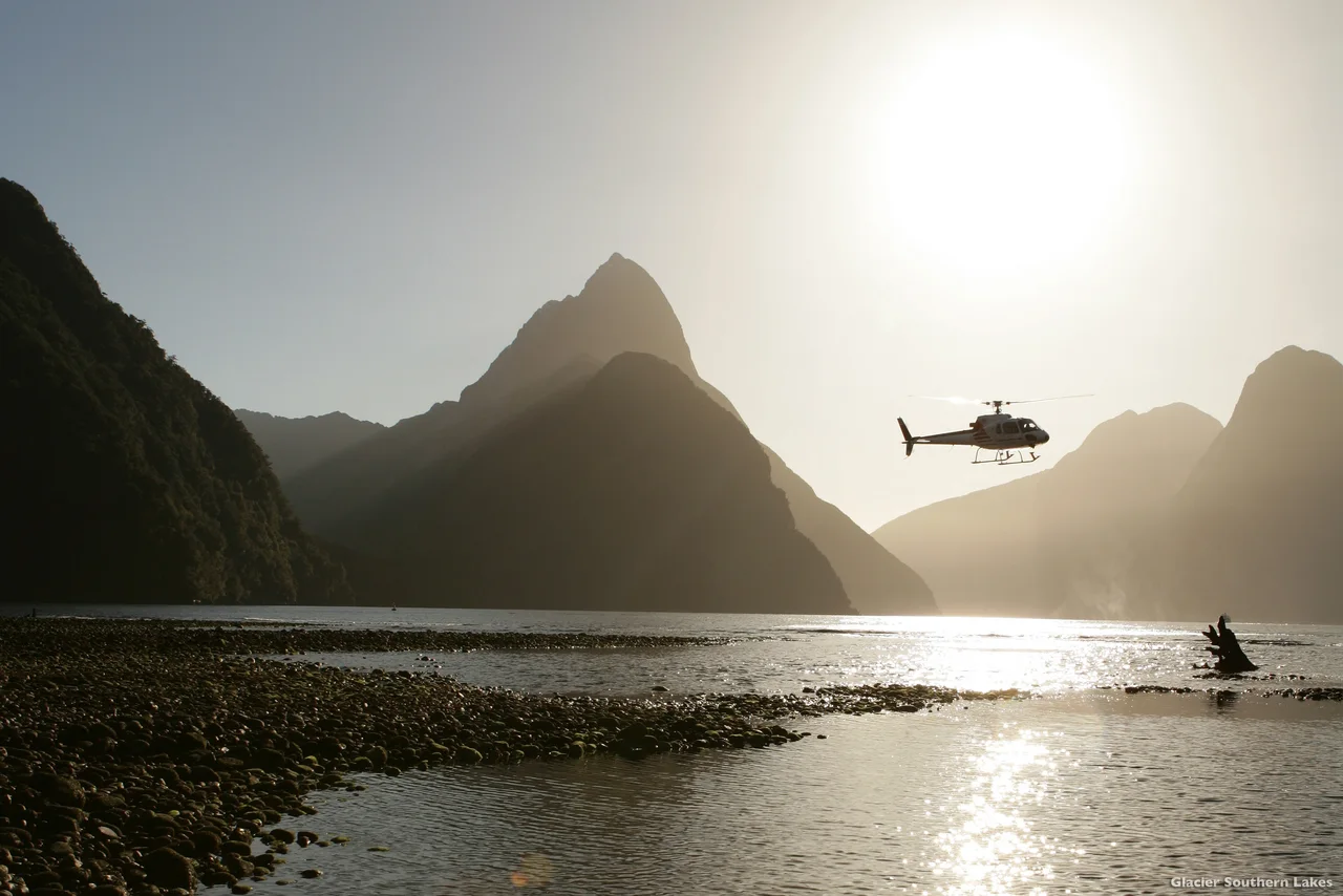 A helicopter flying above a sunlit fjord surrounded by steep mountains.