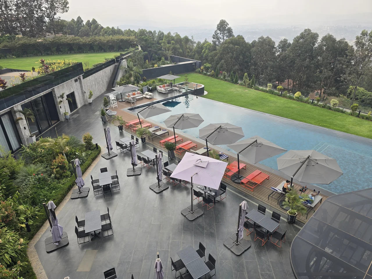A pool and dining terrace with parasols overlooking green gardens and trees.