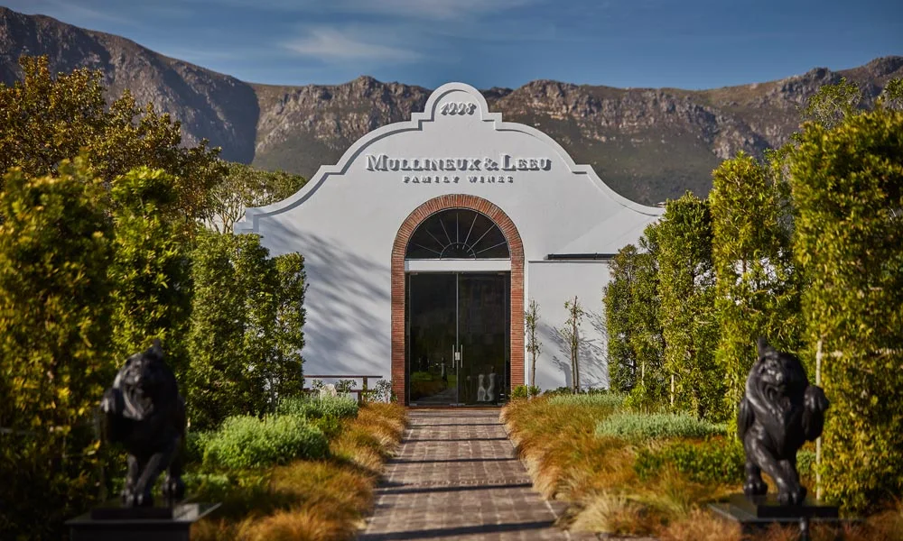 A white winery building framed by trees and mountains.