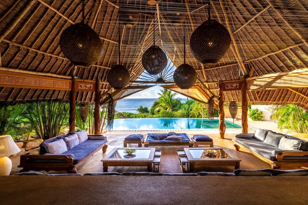 An open-sided villa lounge beneath a thatched roof faces a pool, palm garden and ocean.