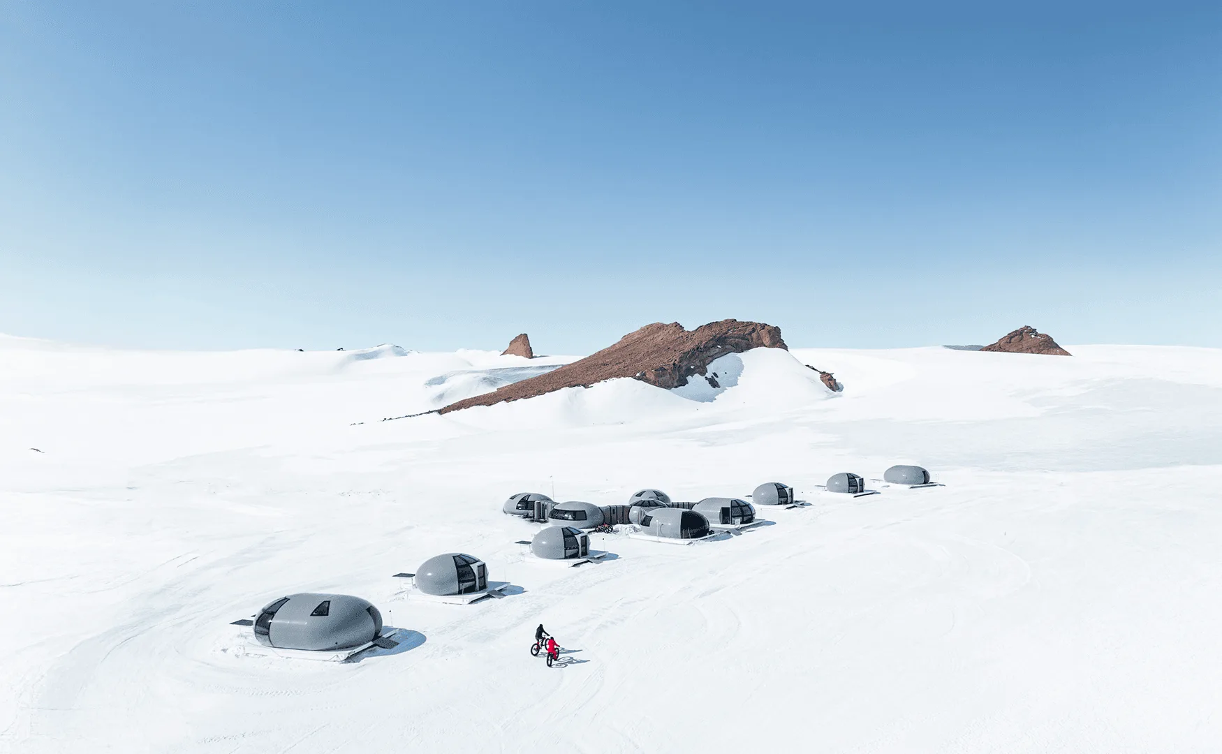 Grey dome-shaped shelters stand on snowy terrain, with one person nearby.