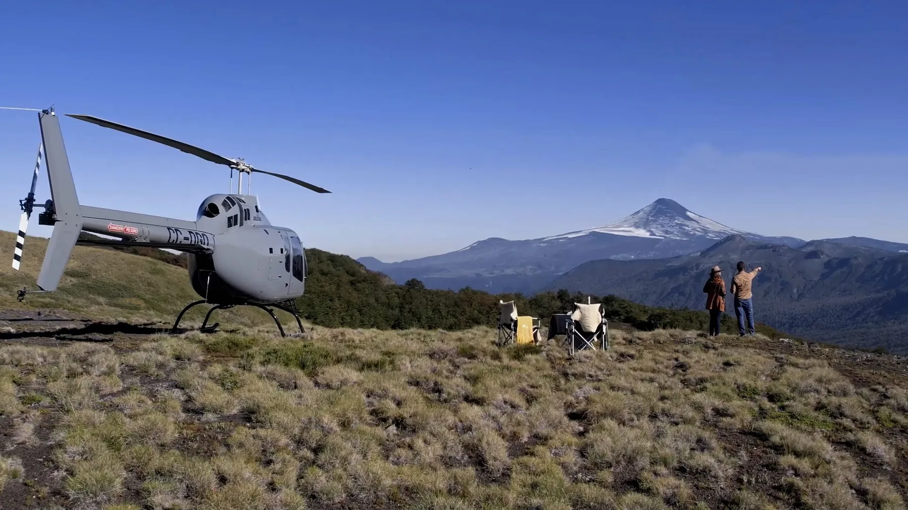 A helicopter stands on a mountain ridge with a volcano in the distance.