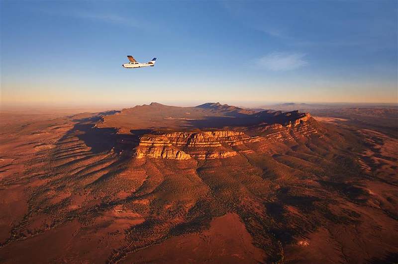 A small aircraft flies above the red ridges of Australia's outback at sunset.