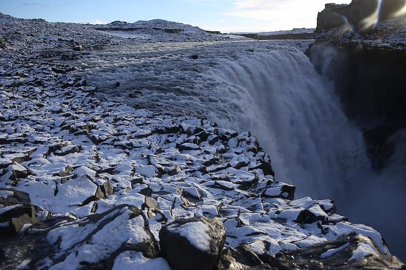 A broad waterfall runs over dark volcanic rock in a wintry Icelandic landscape.