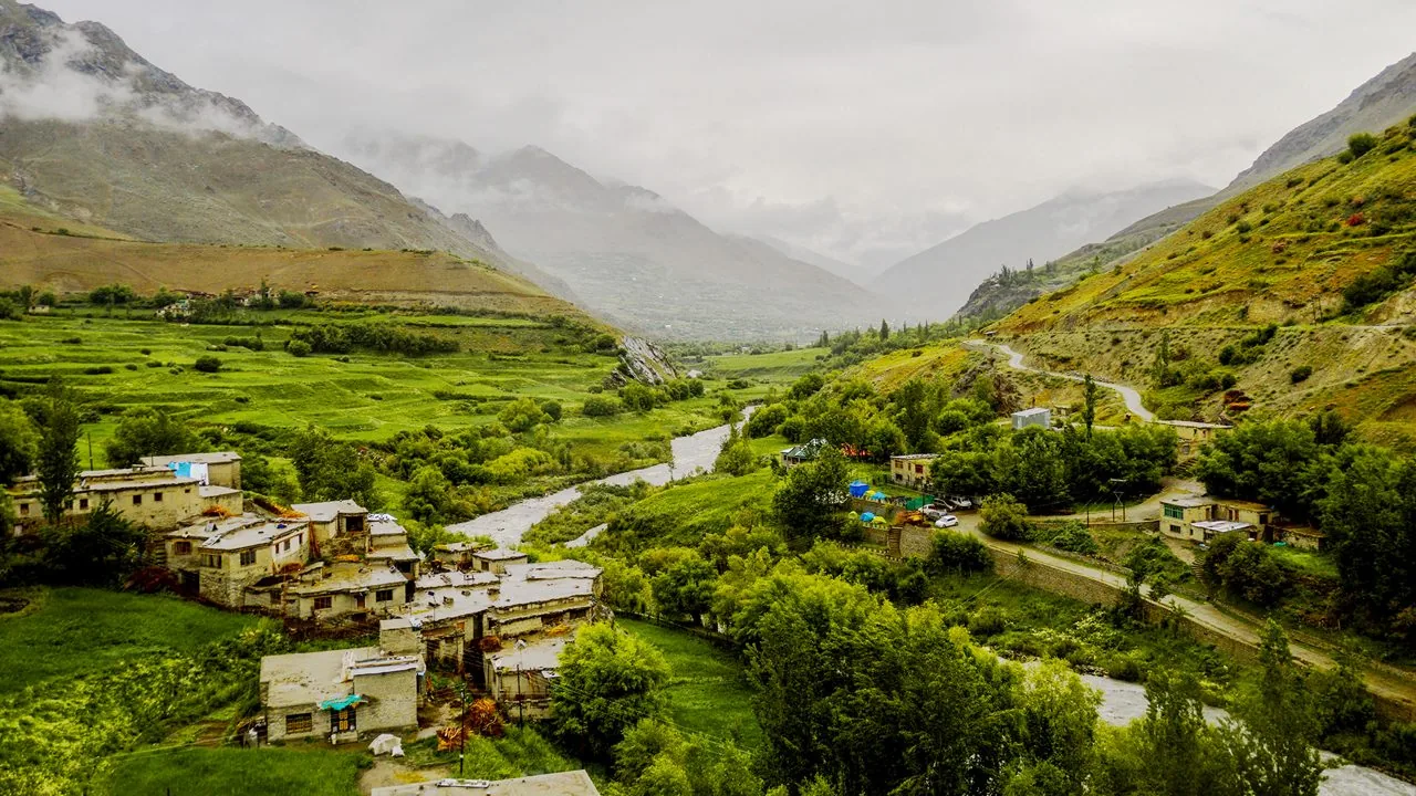 A green Ladakh valley with a stream, scattered homes, and steep mountains.