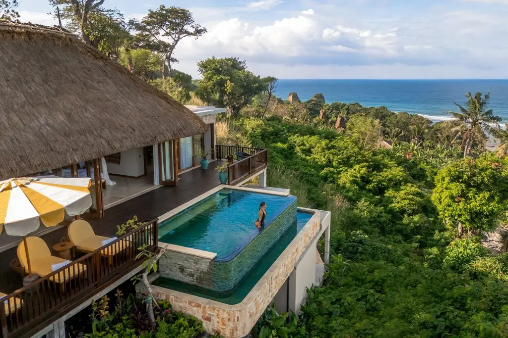 A resort pool and thatched villa overlook lush Sumba vegetation and the ocean.