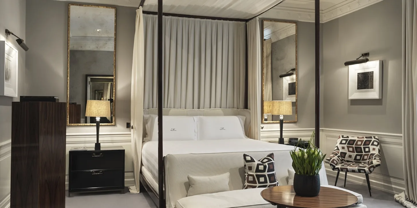 A four-poster bed anchors a refined hotel room with neutral furnishings and pale curtains.